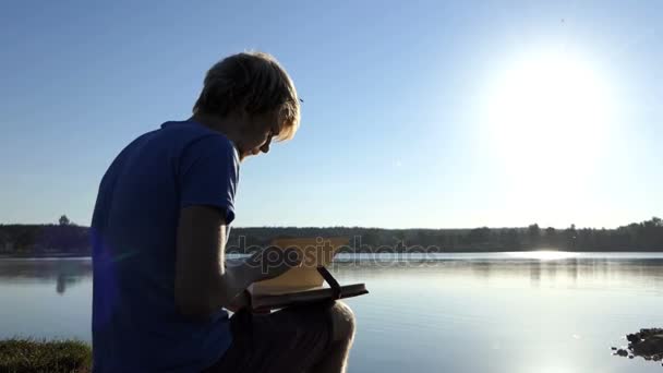 Jovem olha para um álbum de família em um banco de lago no verão — Vídeo de Stock