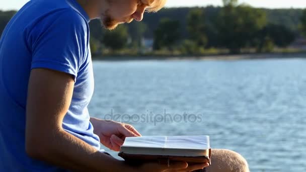Молодой человек смотрит на альбом. На берегу озера летает пчела. — стоковое видео