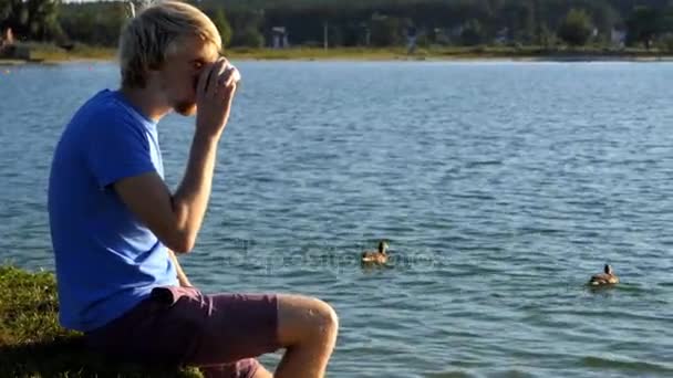 Молодой человек пьет чай на берегу озера с плавающими утками — стоковое видео