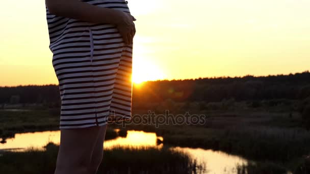 Zwangere vrouw in een gestreepte jurk staat aan de oever van een meer bij zonsondergang — Stockvideo