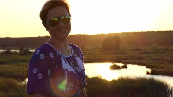 Γυναίκα με γυαλιά ηλίου χαμόγελα σε μια τράπεζα στη λίμνη στο ηλιοβασίλεμα σε slo-mo — Αρχείο Βίντεο