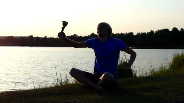 Happy man sits and raises his champion bowl on a lake bank in slo-mo — Stock Video