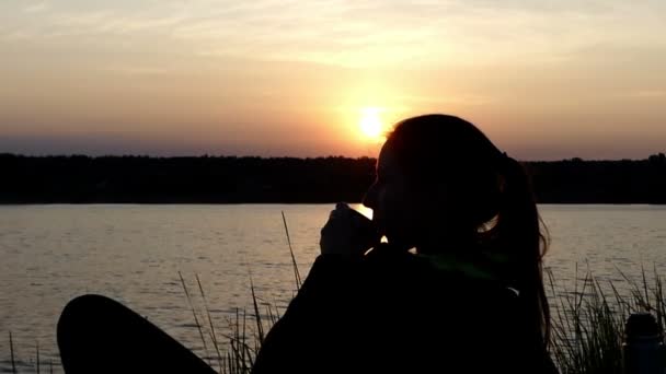 Mujer joven se encuentra en una orilla del lago, bebe té y ríe — Vídeo de stock