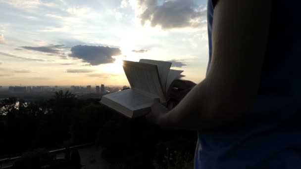 Jovem estudante senta-se e lê um livro em um gramado ao pôr do sol em slo-mo — Vídeo de Stock