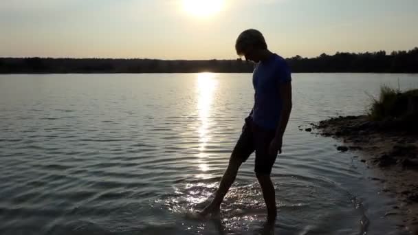 Joven empuja su pie en el agua del lago y lo mueve al atardecer — Vídeos de Stock