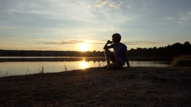 Joven bebe té sentado en una orilla del lago al atardecer en slo-mo — Vídeos de Stock