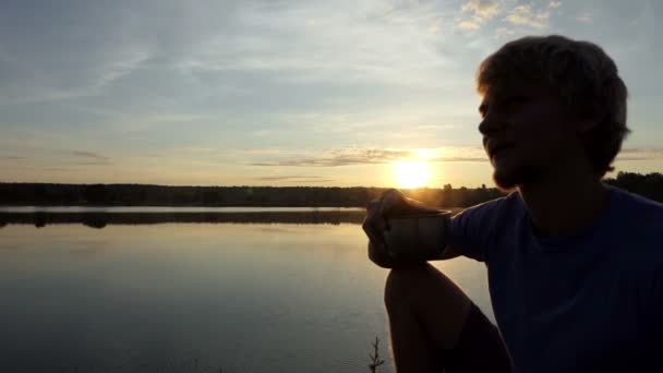 Homem feliz bebe chá sentado em um banco do lago ao pôr do sol em slo-mo — Vídeo de Stock