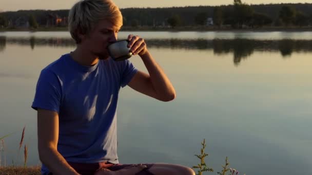 Blond homme boit du café assis sur la rive d'un lac au coucher du soleil à Xo@-@ mo — Video