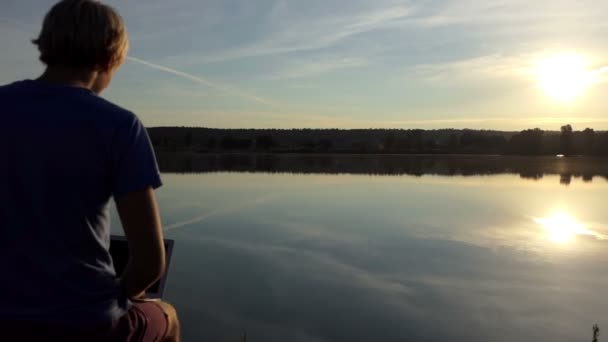 El hombre creativo trabaja con su portátil en una orilla del lago en slo-mo — Vídeo de stock