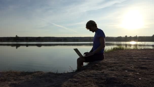 Pro hombre trabaja con su portátil en una orilla del lago en slo-mo — Vídeo de stock