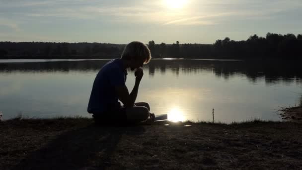 Jonge man leest een boek aan de oever van een meer bij zonsondergang in slo-mo — Stockvideo