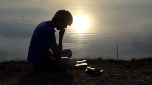 Ο δημιουργικός άνθρωπος διαβάζει ένα βιβλίο σε μια τράπεζα στη λίμνη στο ηλιοβασίλεμα σε slo-mo — Αρχείο Βίντεο