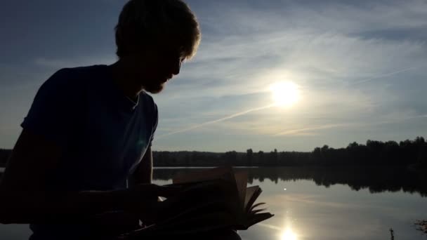 C-student kijkt door zijn boek op de oever van een meer in slo-mo — Stockvideo