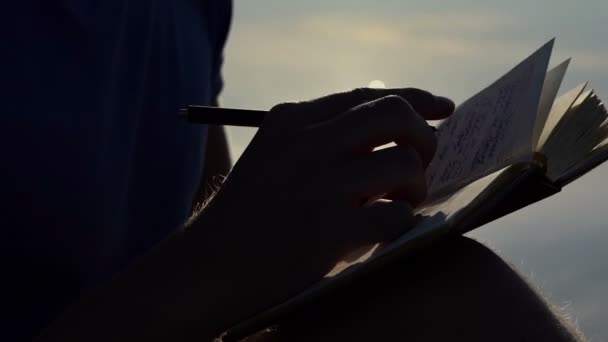 Jonge man schrijft iets in zijn Kladblok bij zonsondergang in slo-mo — Stockvideo