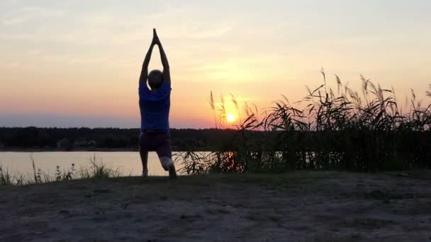 Young man practices yoga on a lake bank at sunset in summer — Stock Video