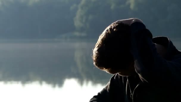 Joven se sienta en una orilla del lago y se frota el cuello — Vídeo de stock