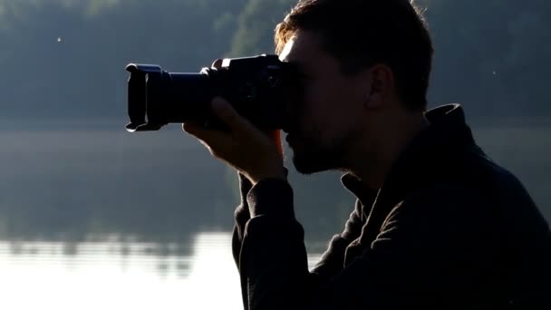 Young man shoots a nice lake with his camera at sunset — Stock Video