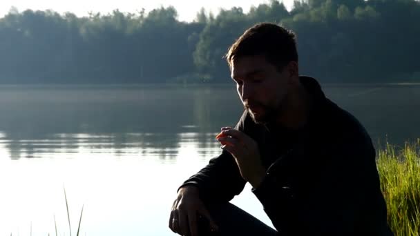 Νεαρός άνδρας κάθεται σε μια τράπεζα στη λίμνη και τρώει ένα καρότο — Αρχείο Βίντεο