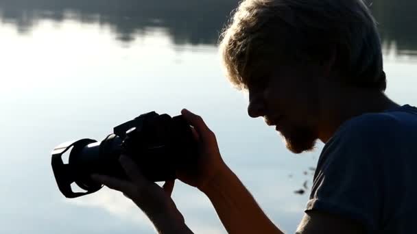 Homme blond assis à un lac de forêt et regarde son appareil photo en été — Video