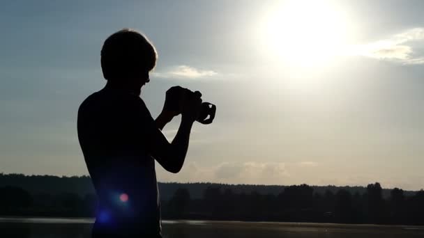 Blonde man staat op een bos meer en lijkt op zijn camera in zomer — Stockvideo