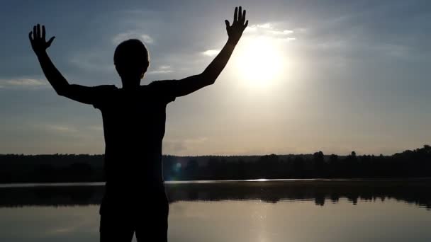 Joven levanta las manos felizmente en una orilla del lago al atardecer en cámara lenta — Vídeos de Stock