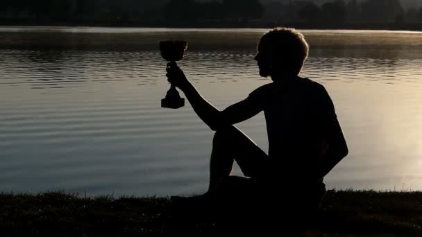 Young man sits on a lake bank and looks at a winner bowl in slo-mo — Stock Video