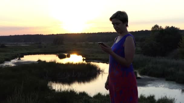 Mujer agradable llama en una orilla del lago al atardecer — Vídeos de Stock