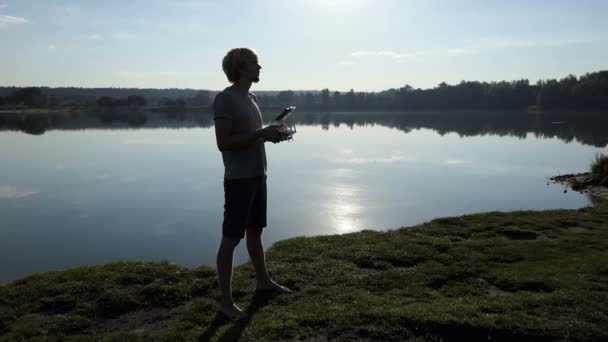 Jovem olha para cima e opera um drone em um banco de lago em slo-mo — Vídeo de Stock