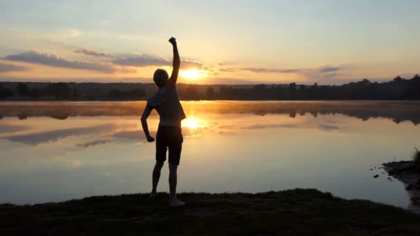 Joven levanta el puño sobre un lago en una espléndida puesta de sol en slo-mo — Vídeos de Stock