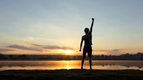 Homem independente levanta o punho sobre um lago ao pôr-do-sol em Slo-mo — Vídeo de Stock