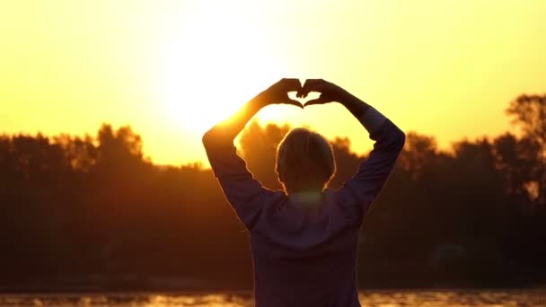 Jonge man houdt een gebaar van liefde op de oever van een meer bij zonsondergang — Stockvideo