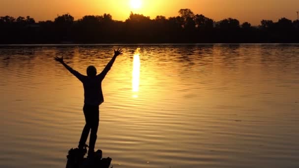 Hombre alegre levanta dos manos en una orilla del lago al atardecer en slo-mo — Vídeo de stock