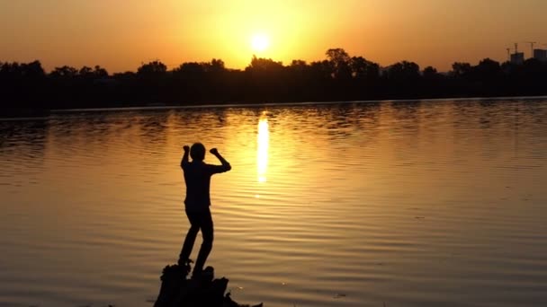 Gelukkig man werpt twee handen op de oever van een meer bij zonsondergang in slo-mo — Stockvideo