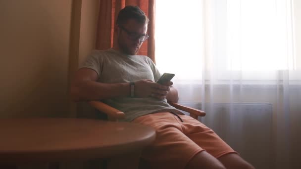 Bir adam otel odasının penceresinin yanındaki sandalyeye oturur ve telefonla çalışır.. — Stok video