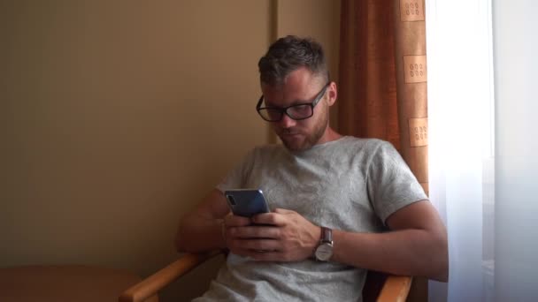 Hombre con gafas, pelo rojo, reloj se sienta en la silla y escribe su teléfono . — Vídeos de Stock