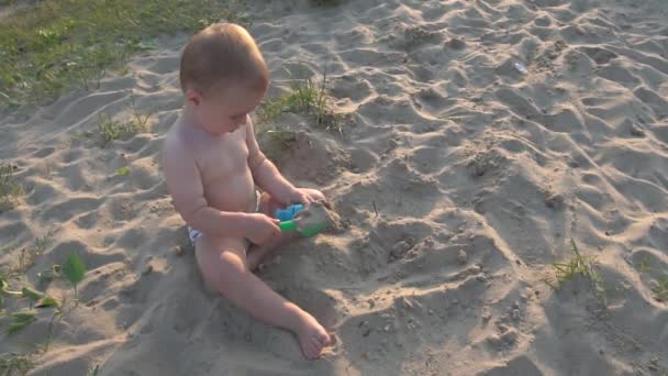 Niño pequeño se sienta en la arena y juega con un cubo y una pala . — Vídeos de Stock