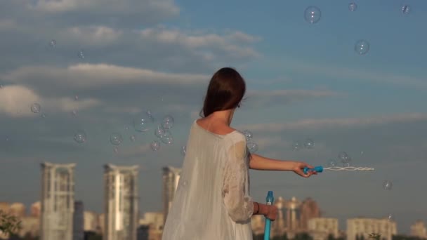 Una jovencita agita la mano y sopla burbujas de jabón en el fondo de la ciudad . — Vídeos de Stock
