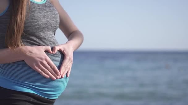 Jonge vrouw tonen hart op haar zwanger buik op zee kust — Stockvideo