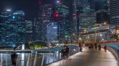 Yürüyen insanlarla Esplanade Köprüsü ve arka planda merkez gökdelenleri olan Singapur gece zaman çizelgesi. Aydınlatılmış kuleler suya yansıyor