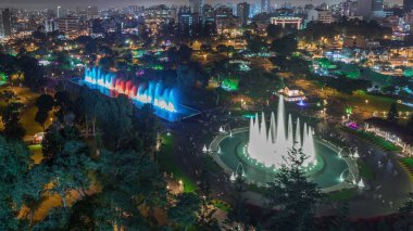 Havadan Park of the Reserve 'e en büyük çeşme kompleksiyle gece vakti aydınlatılmış renkli sihirli su devresini gösteren panoramik bir görüntü. Yeşil çimenler ve ağaçlar. Arkaplanda şehir silüeti