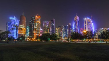Geceleri Doha 'nın ufuk çizgisi. Yıldızlı gökyüzü Park Timelapse, Katar' dan görülüyor. Ön planda ağaçlar. Aydınlatılmış gökdelenler ve kuleler