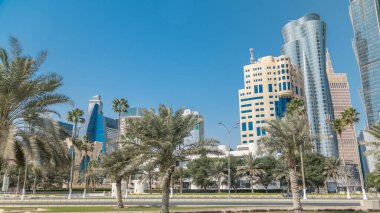 Doha 'nın ufuk çizgisi Park Timelapse, Katar' dan görüldü. Ön planda ağaçlar ve palmiyeler. Arka planda Mordern gökdelenleri ve kuleleri. Yolda trafik vardı.