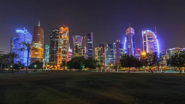 Geceleri Doha 'nın ufuk çizgisi. Yıldızlı gökyüzü Park Timelapse, Katar' dan görülüyor. Ön planda ağaçlar. Aydınlatılmış gökdelenler ve kuleler