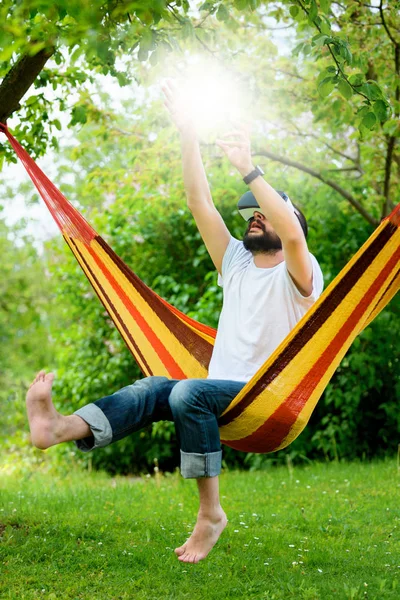 Jovem barbudo vestindo óculos de realidade virtual relaxando em uma rede de jardim. Estilo de vida VR divertido e relaxe conceito — Fotografia de Stock