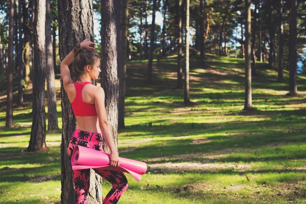 Jovem mulher atlética ajuste vestindo relógio inteligente e segurando tapete de ioga, pronto para seu treino em uma floresta. Conceito de felicidade mental e corporal — Fotografia de Stock