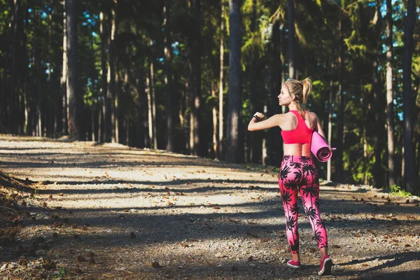 Молодая спортивная женщина в лесу в умных часах и с ковриком для йоги, уходя от камеры. Разум и тело понятие счастья — стоковое фото