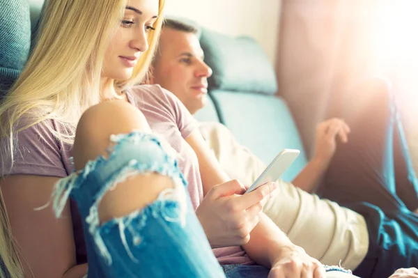 Unga snygga paret sitter på en avkopplande soffa i deras moderna hem. Kvinna som håller mobiltelefon, surfa på webben, mannen framför TV — Stockfoto