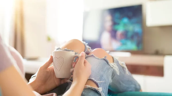 Vacker ung kvinna avkopplande i soffan hemma, titta på tv och njuta av kaffe — Stockfoto