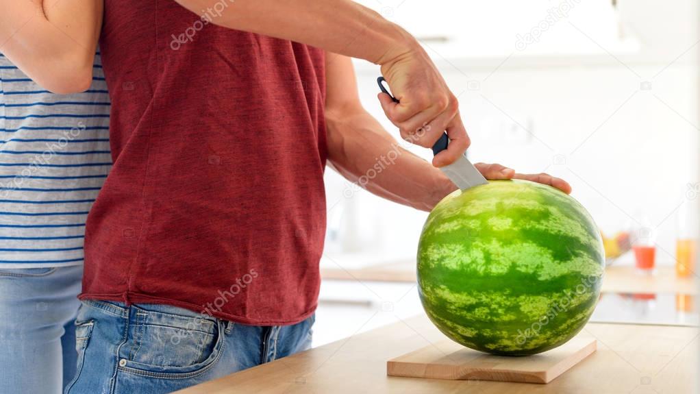Young couple in a kitchen slicing red watermelon. Couple in their large contemporary white kitchen