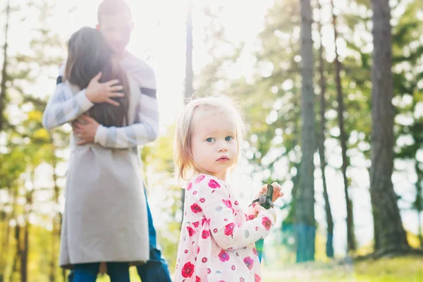 Gelukkige jonge familie nemen van een wandeling in een park. Ouders knuffelen in de achtergrond met peuter meisje dochter kijken camera op de voorgrond. — Stockfoto